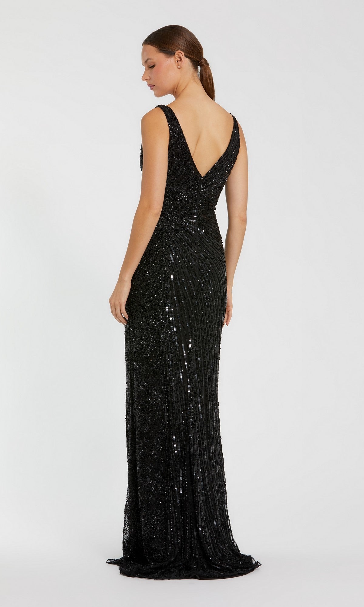 Woman in a sleeveless, beaded Mac Duggal 5473 formal dress with a fitted, floor-length black silhouette and deep V-back, stands against a plain white background facing away from the camera.